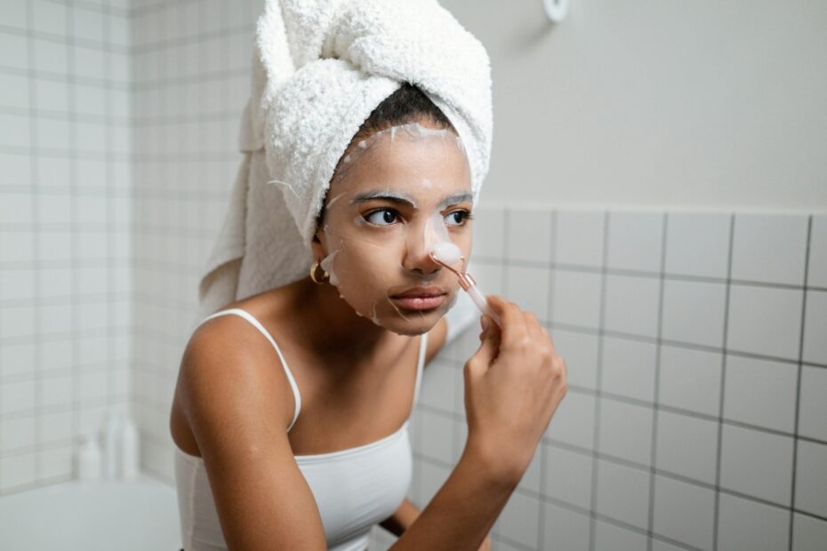 mulher negra com cabelo enrolado na toalha, olhando para o espelho e colocando máscara facial